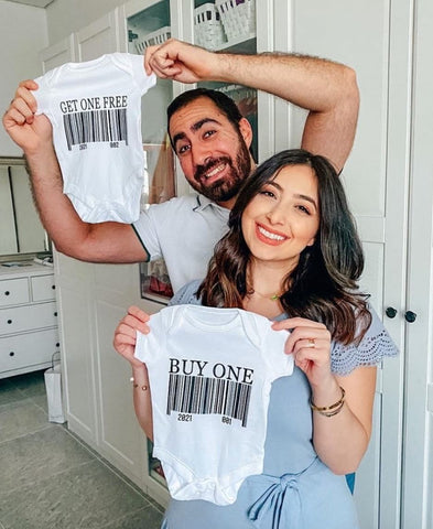 A couple holds baby onesies that say "Buy One" and "Get One Free" to announce a twin pregnancy