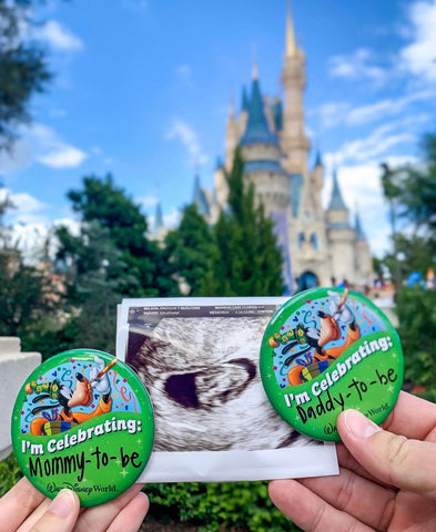 A disney pregnancy announcement photo: Hands hold a sonogram and Disney pins that say "Celebrating Mommy to Be" and "Celebrating Daddy to Be"