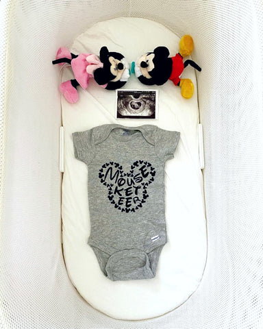 Disney pregnancy announcement photo of a Mouse ears onesie and Mickey Mouse dolls in a SNOO bassinet