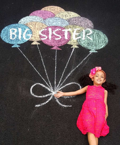 DIY pregnancy announcement photo of a little girl laying on asphalt holding a chalk-drawn bunch of balloons that say "BIG SISTER"