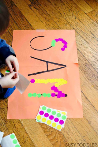 Toddler uses dot stickers to trace the name SAM on a piece of paper.