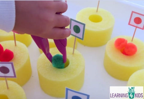 A toddler uses plastic tongs to put pom-poms into the donut-shaped pieces of a cut-up pool noodle.