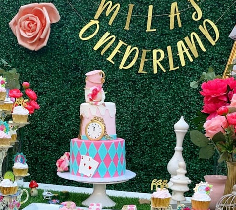 Alice in Wonderland-themed first birthday cake on a table with a banner in the background reading “Mila’s Onderland”