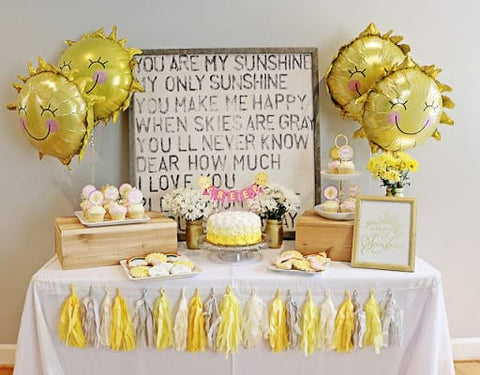 First birthday party table displaying sun-shaped mylar balloons, a yellow cake, and sun, cloud, and rainbow cookies. 