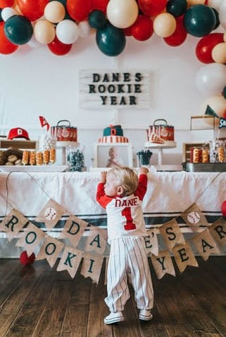 The back of a toddler boy wearing a baseball uniform standing in front of a baseball-themed party table.