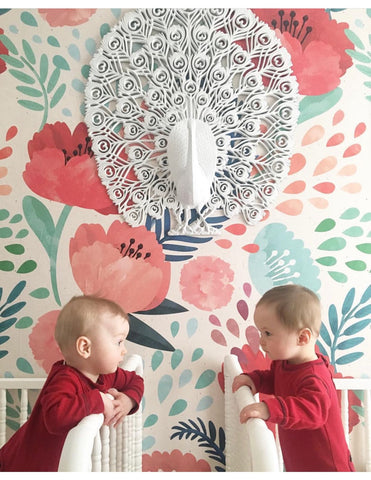 Twin babies look at each other from their cribs, which are placed against a poppy wallpaper