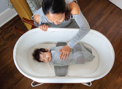 A mother gently touches the belly of her baby, who is sleeping in a SNOO bassinet.