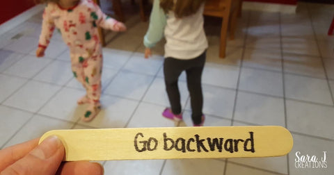 Hand holding a popsicle stick with the words "go backward" written on it as two toddlers stand in background.