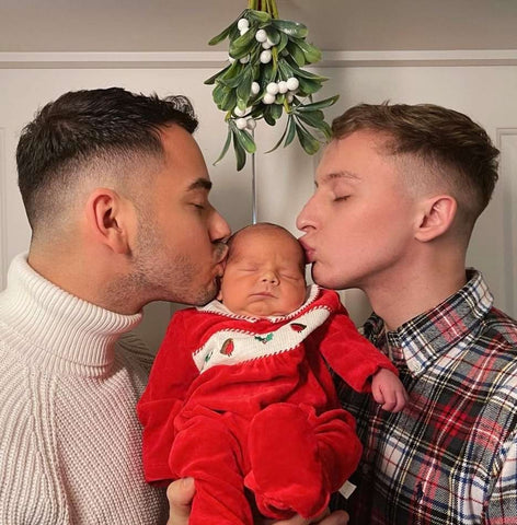 Two dads kiss their baby's cheeks under the mistletoe to celebrate baby's first holiday