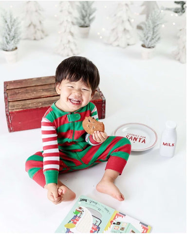 A little boy poses in Christmas PJs near milk and cookies for a holiday photo