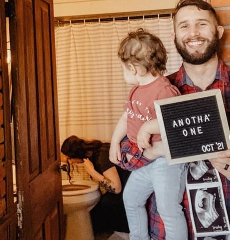 Dad holding ultrasound and letterboard in foreground as mom pukes in toilet in background