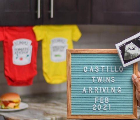 Baby onesies made to look like ketchup and mustard bottles displayed behind letterboard announcing a twin pregnancy