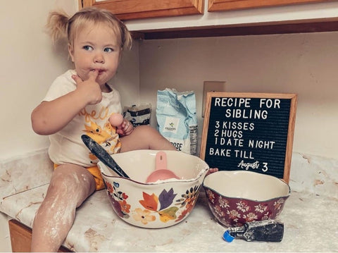 A toddler poses in the kitchen next to a letterboard pregnancy announcement that says "recipe for sibling'"