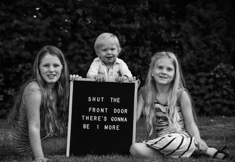 Three siblings pose with a letterboard pregnancy announcement that reads "shut the front door there's going to be 1 more"