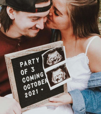 A couple poses with a letterboard pregnancy announcement that says "party of 3 coming October"