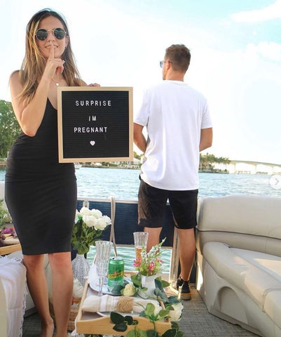 A woman poses with a letterboard pregnancy announcement that says "Surprise I'm pregnant" while her partner stands in the background with his back turned
