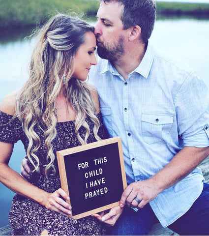 A couple poses with a pregnancy announcement letterboard that says "for this child I have prayed"