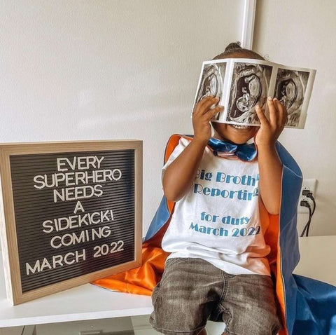 A toddler in a cape poses next to a letterboard pregnancy announcement that says "every superhero needs a sidekick"