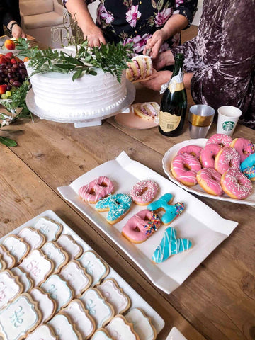 White gender reveal cake displayed on table with pink and blue donuts and cookies.