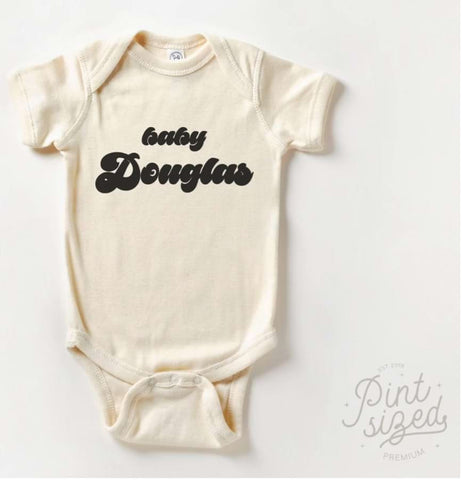 Pregnancy announcement onesie that says "Baby Douglas" in 70s-style text