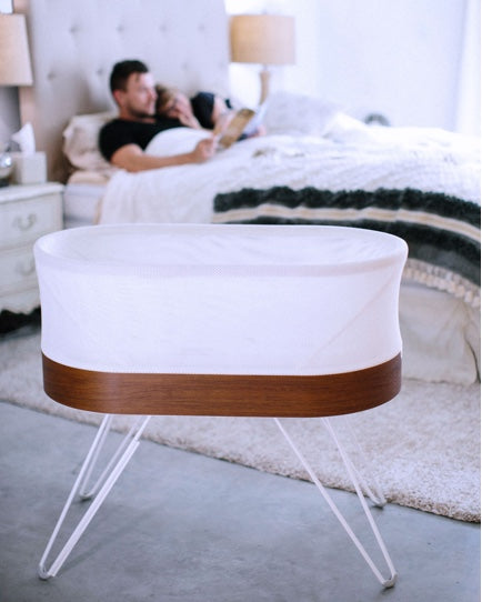 A SNOO bassinet sits in the foreground of a master bedroom, while a couple snuggles on the bed in the background.