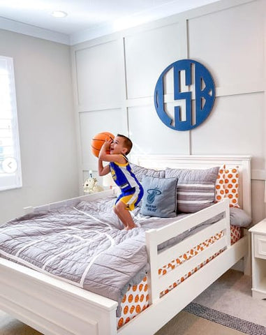 Toddler stands on a bed with basketball-printed sheets, poised to shoot a basketball.