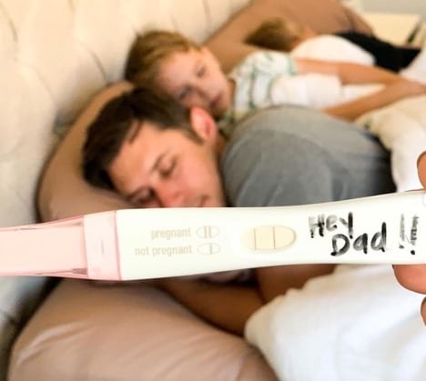 Positive pregnancy test with "Hi Dad!" written on it held in front of a sleeping father