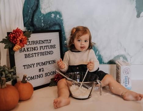 A toddler poses with baking equipment and a letterboard announcing Baby #2