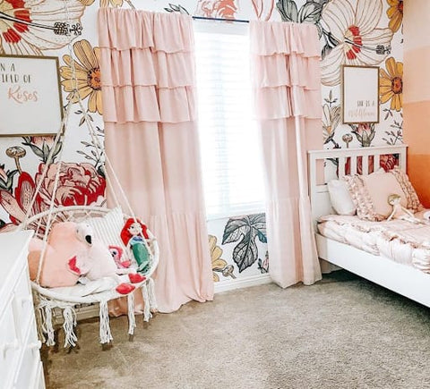 Toddler bedroom with floral print wallpaper, pink curtains, and a white wicker swing hanging from the ceiling. 