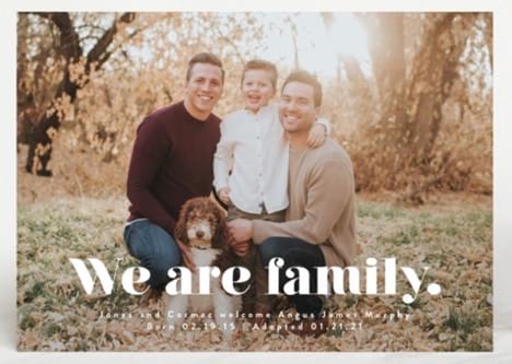 Two dads and adopted son pose on card that says "we are family" to announce adoption