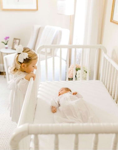 A toddler peeks at a baby sleeping in a vintage crib in an all-white nursery