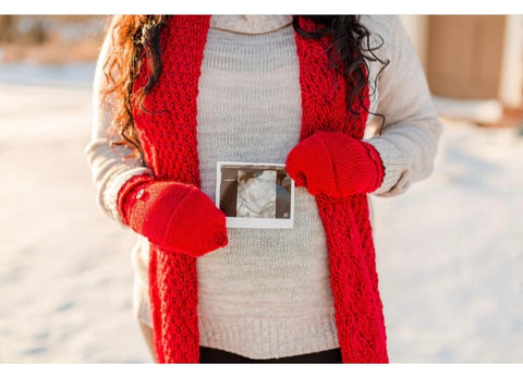 An expecting mom wears red knit accessories and holds a sonogram photo to announce a pregnancy