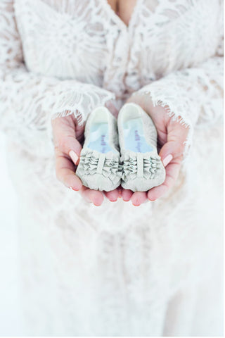 A mom-to-be wears a white gown and holds white booties in a winter pregnancy announcement