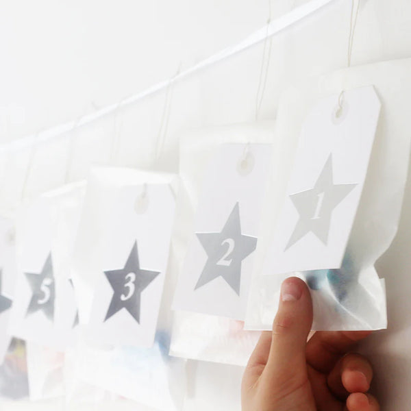 white parcel tags have been embossed with one central silver star and each star is numbered 1-25. A hand holds a small glassine envelope behind the number 1, there is obviously something in the envelope