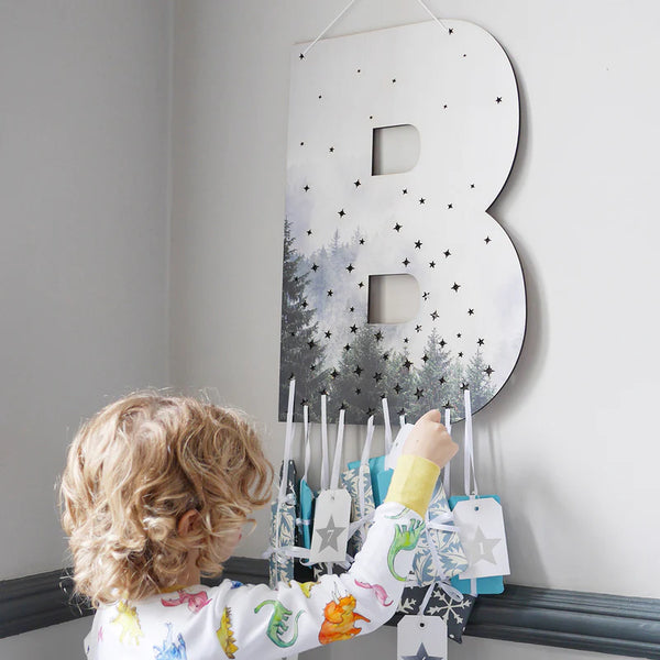 A large letter B, cut from wood and painted with a forest scene, has star shaped holes in it. A small boy in pyjamas is reaching out for one of the gifts that has been strung through a hole.