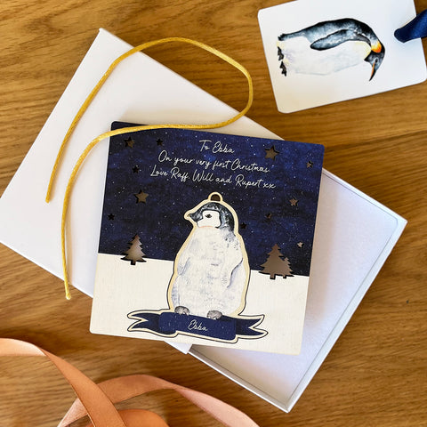 baby emperor penguin chick, wooden Christmas tree decoration and card. Sitting on top of a white gift box