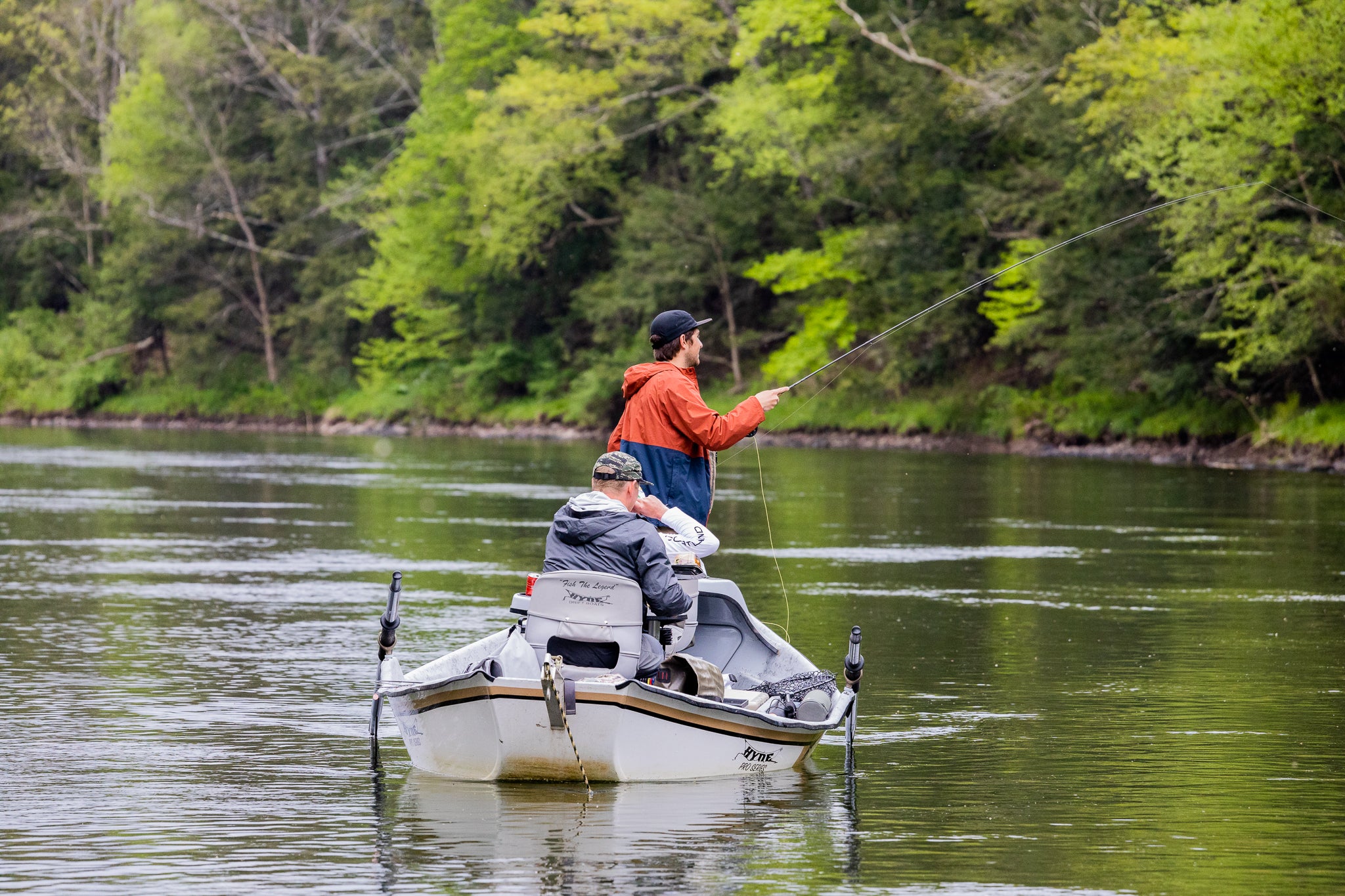 Three guys fishing on a boat. One is standing up fishing, while the other two are sitting down