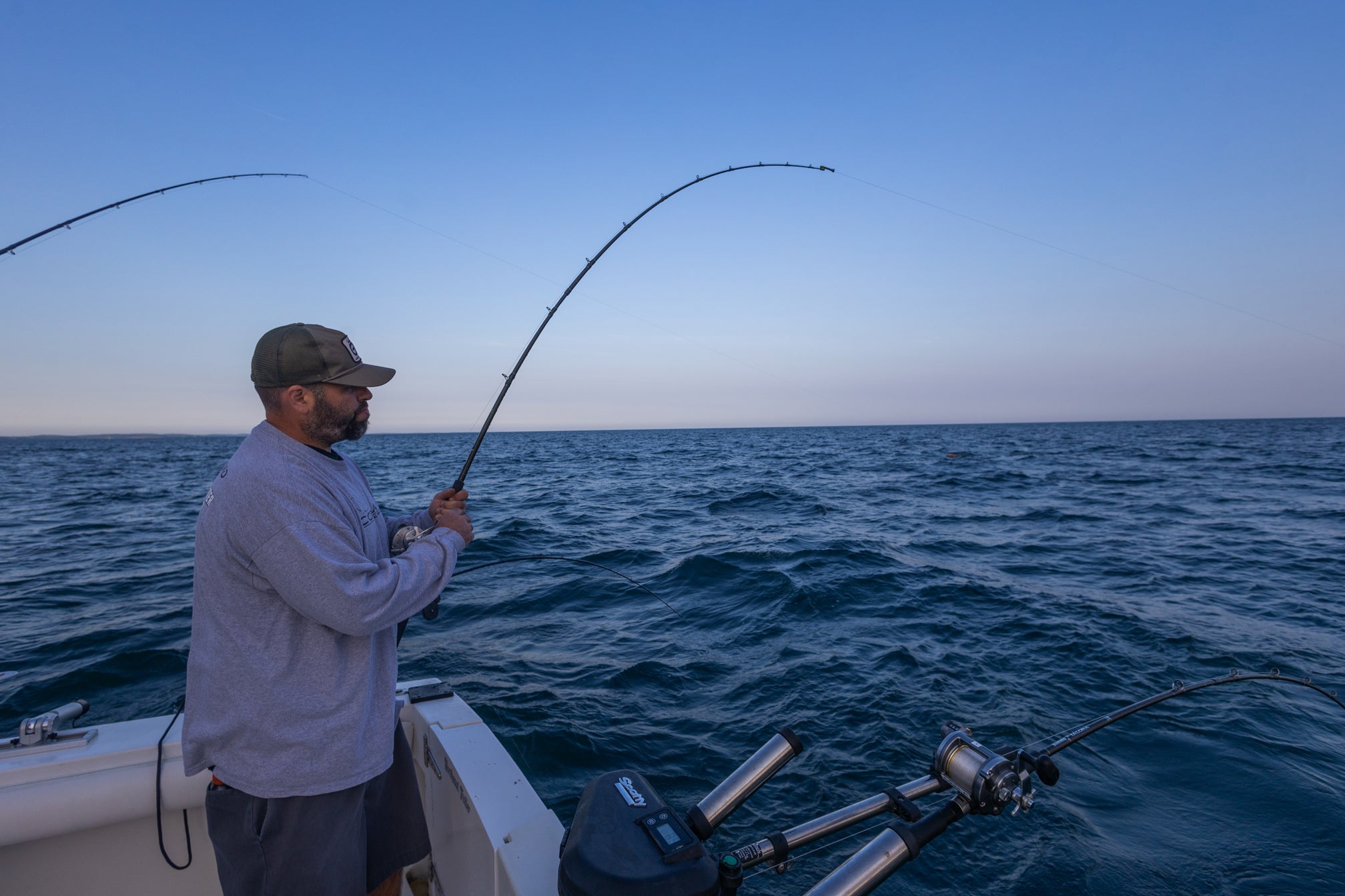 Ein Mann steht auf einem Boot und hält seine Fliegenfischerrute fest. Es gibt eine weitere Fliegenrute, die hinten in einer Halterung gehalten wird