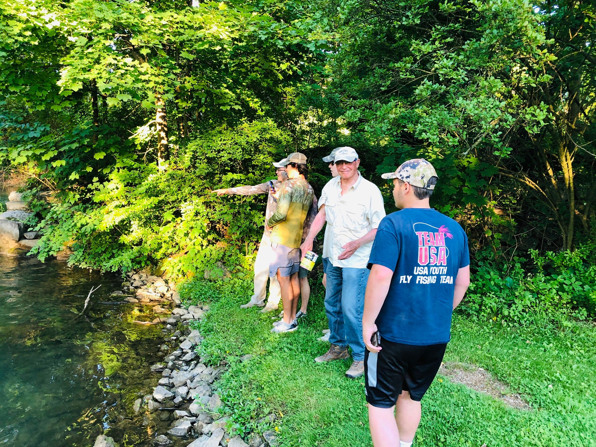 Five people standing on a bank of a river. One is pointing at the water, and another is smiling at the camera.