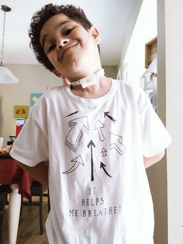 Isaac usando su camiseta It Helps Me Breathe que tiene un montón de flechas apuntando hacia su traqueostomía o traqueotomía.