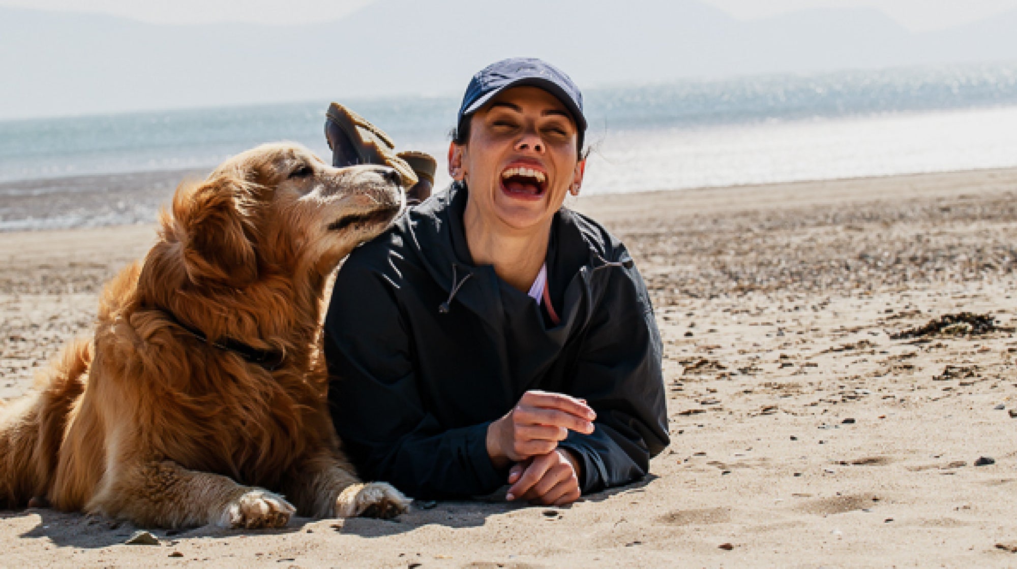 A woman lay on the beach with her golden retriever dog, wearing a Showerproof Popover from ACAI