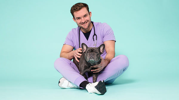 An image of Vet Alex, a 30-something year old man with dark hair, smiling at the camera, wearing lilac vet scrubs, with a grey French Bulldog, against a pale blue background