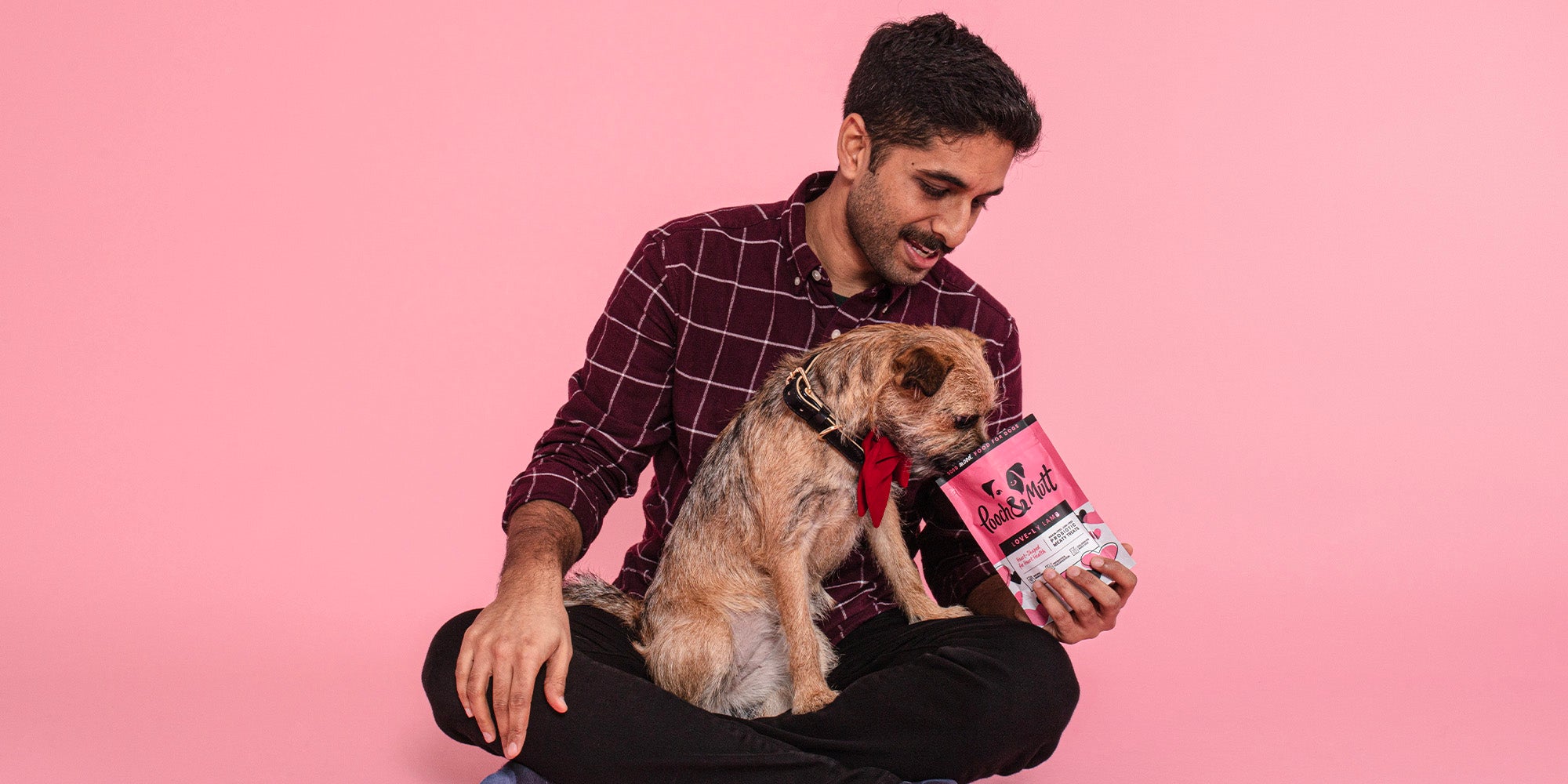 A dark-haired man, feeding his golden coloured dog Pooch & Mutt treats from the bag, against a pale pink background