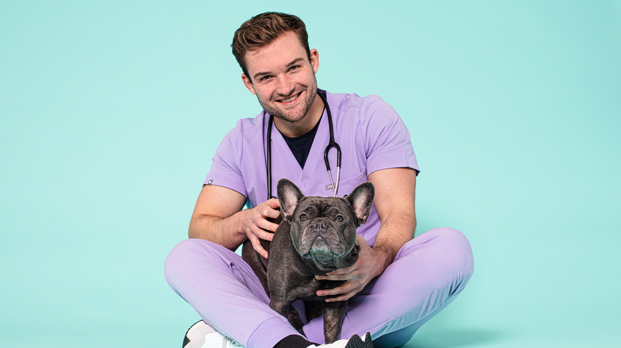 Vet Alex - our Pooch & Mutt Vet. Wearing lilac scrubs, with a grey French Bulldog on his lap, against an aqua coloured background