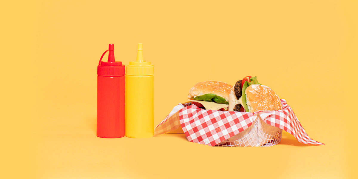 A burger in a wicker basket with a red and white gingham napkin, next to plastic condiment bottles, against a pale orange background