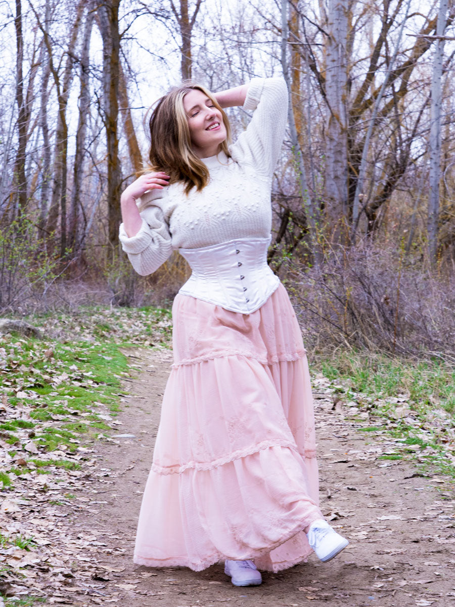 Caylyn in white satin hourglass curve CS-201 waspie underbust corset top worn with flowy pink skirt and white sweater in the woods