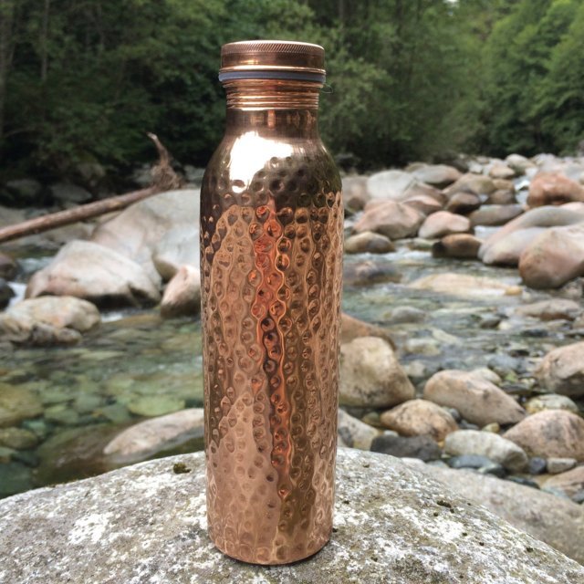 Copper H2O hammered copper water bottle placed on a smooth rock near a river