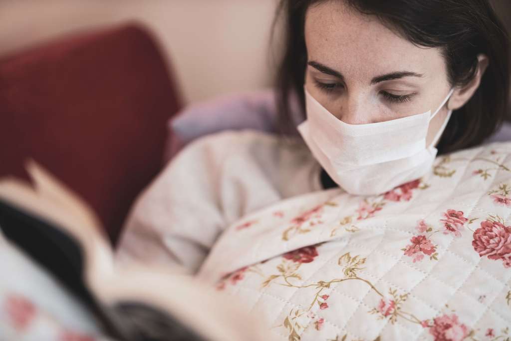 woman wearing surgical mask covered in floral blanket reading a book