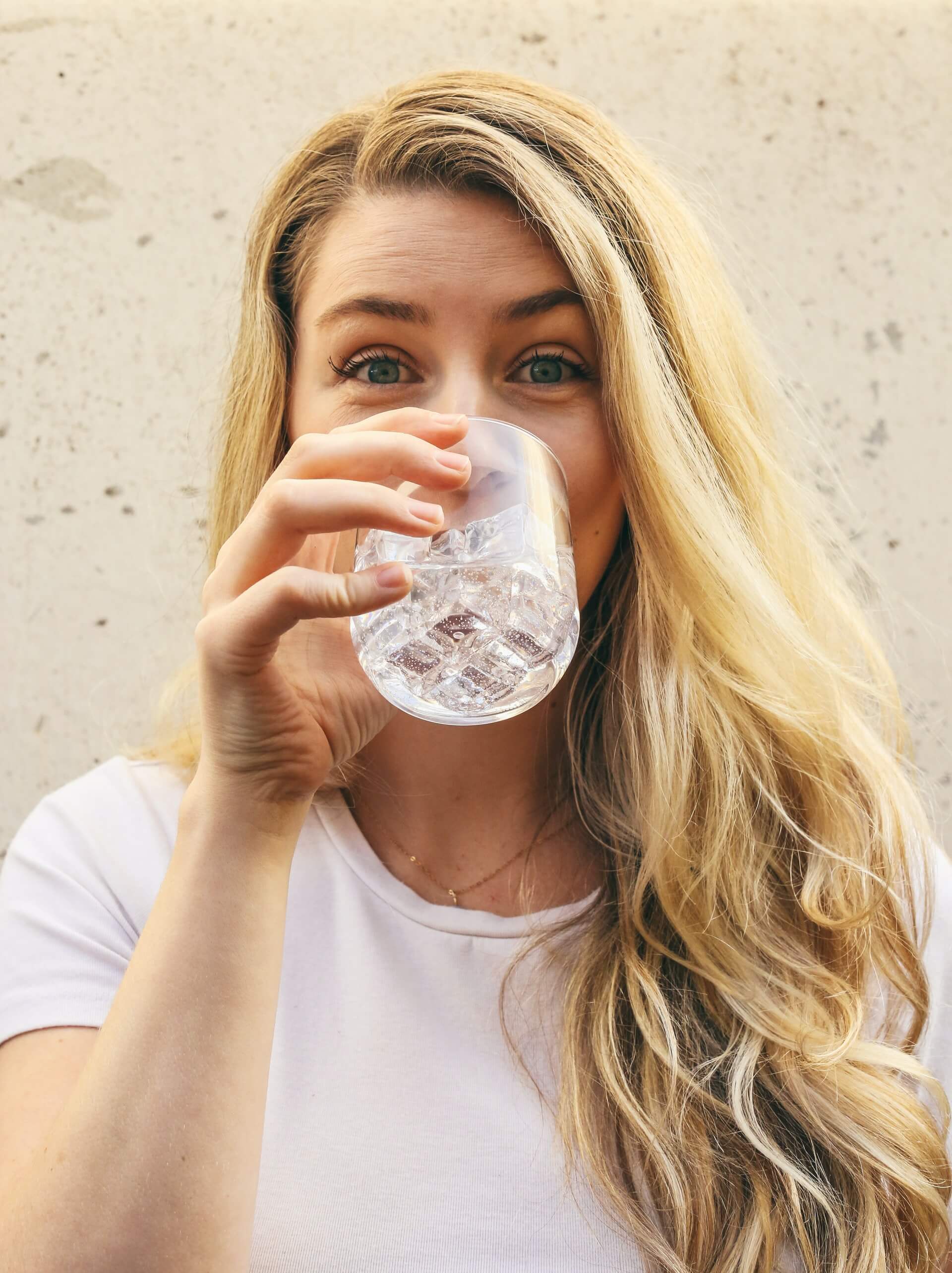 woman blond hair wearing white top drinking water with ice cubes from glass