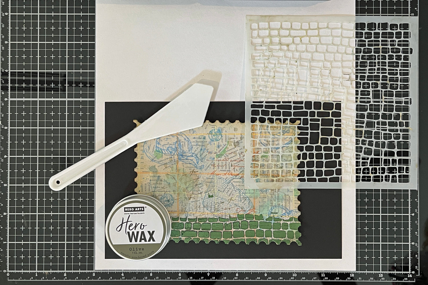 Stenciling a road with Olive Green Hero Wax and a Cobblestone Stencil on a mixed media card background.